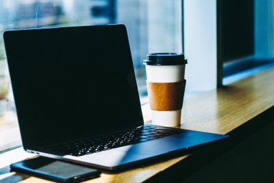 Ноутбук з чорним екраном, що сидить на барі біля вікна, з чашкою кави та смартфоном біля нього.